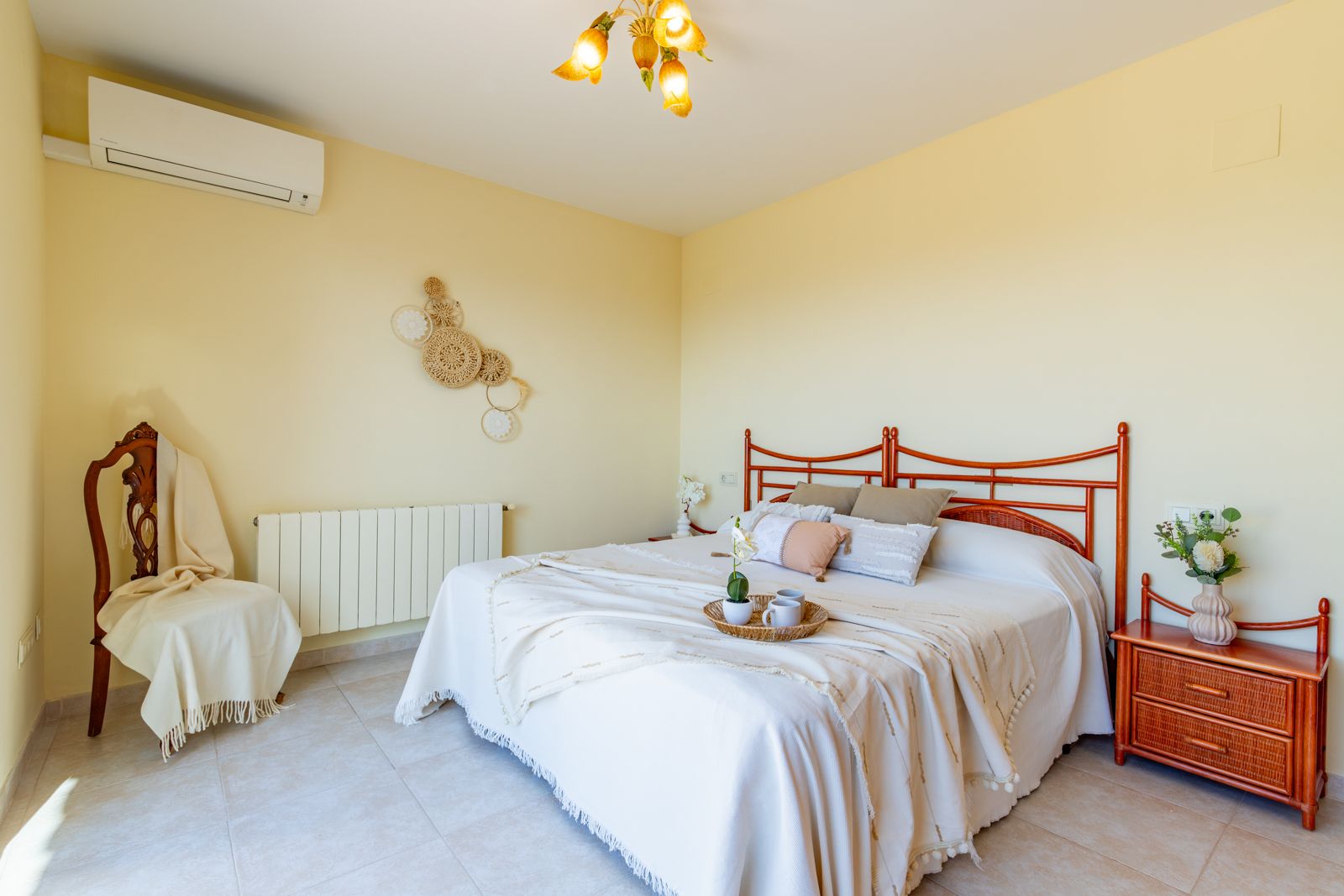 Lichte slaapkamer met tweepersoonsbed, bewerkt houten hoofdbord, bijpassend nachtkastje, split-airconditioning, radiator en neutrale tegelvloer.