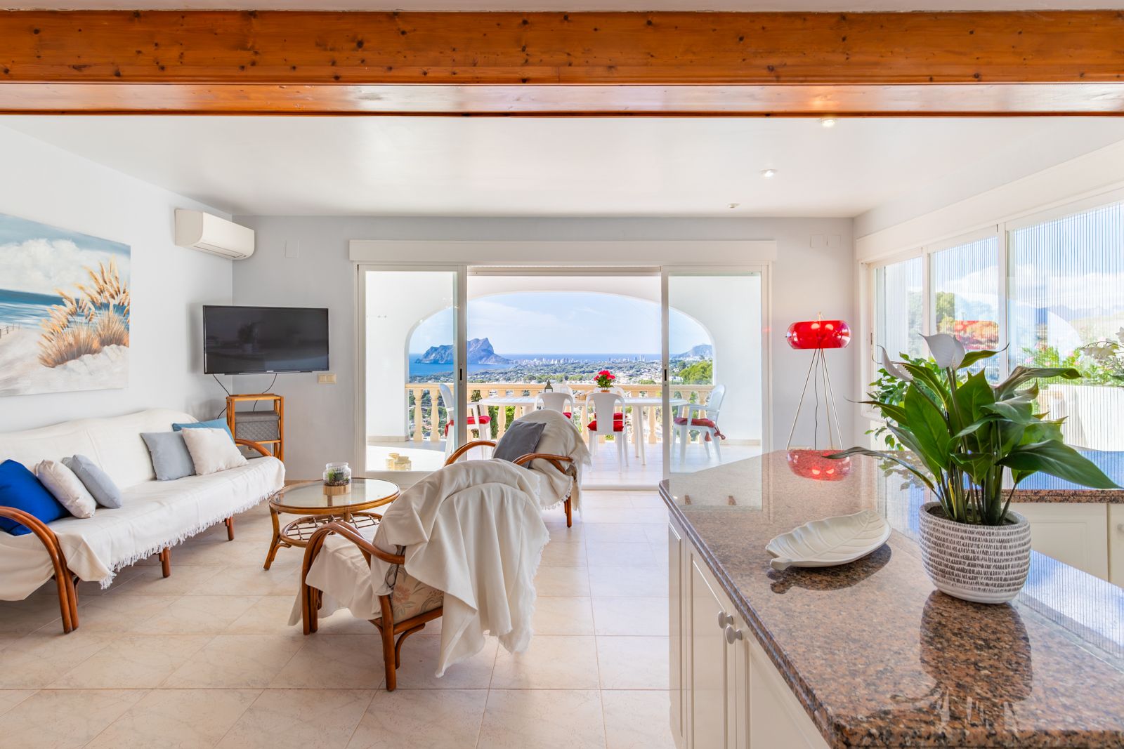 Lichte woonkamer met houten balkenplafond, granieten keukenblad en schuifpui naar het terras met panoramisch uitzicht op de zee en de Peñón de Ifach.