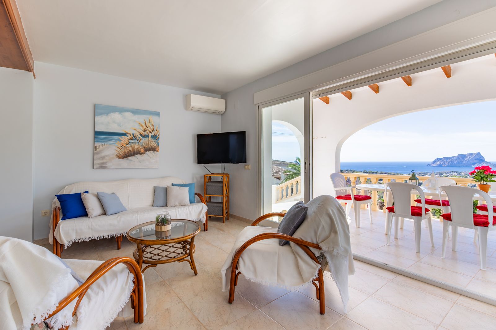 Lichte woonkamer met rotan meubilair en schuifpui naar een overdekt terras. Biedt een panoramisch uitzicht op de Middellandse Zee en de Peñón de Ifach.