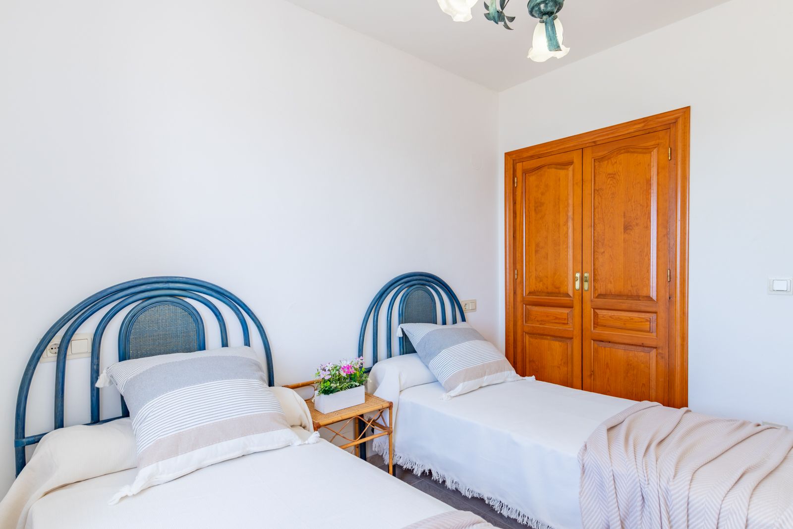 Slaapkamer met twee eenpersoonsbedden, blauwe rieten hoofdeinden en een ingebouwde houten kledingkast. Lichte ruimte met witte muren en een tegelvloer.