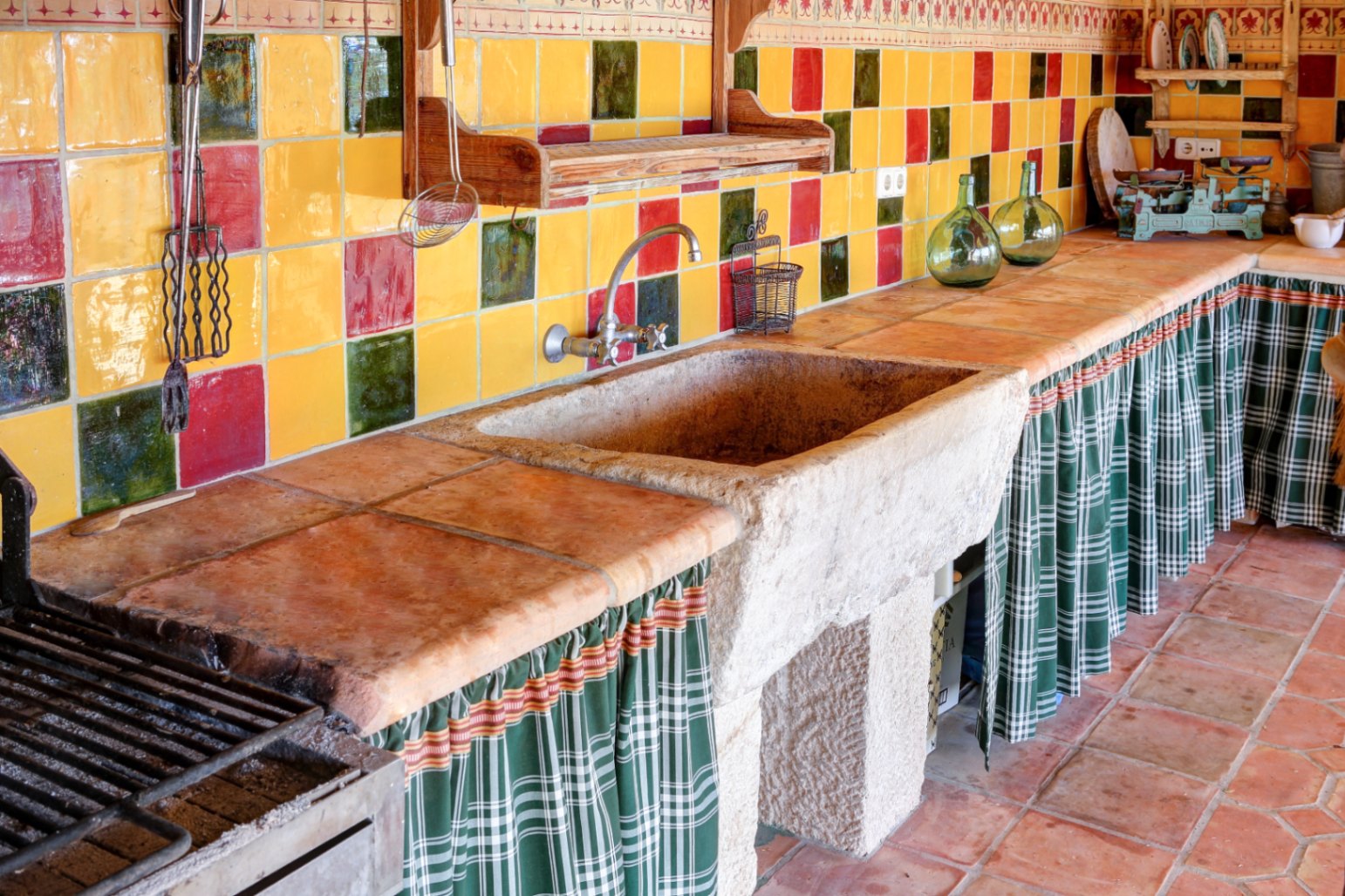 Buitenkeuken met een massief stenen gootsteen, terracotta werkbladen en een kleurrijke keramische achterwand. Voorzien van een barbecuezone en stoffen gordijnen onder het aanrecht.