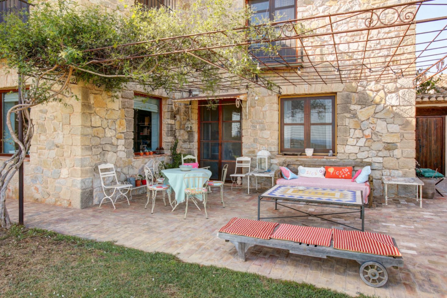 Buitenterras met natuurstenen gevel, terracotta vloer en een smeedijzeren pergola. De ruimte is voorzien van rustiek meubilair en een salontafel met een blad van mozaïek.