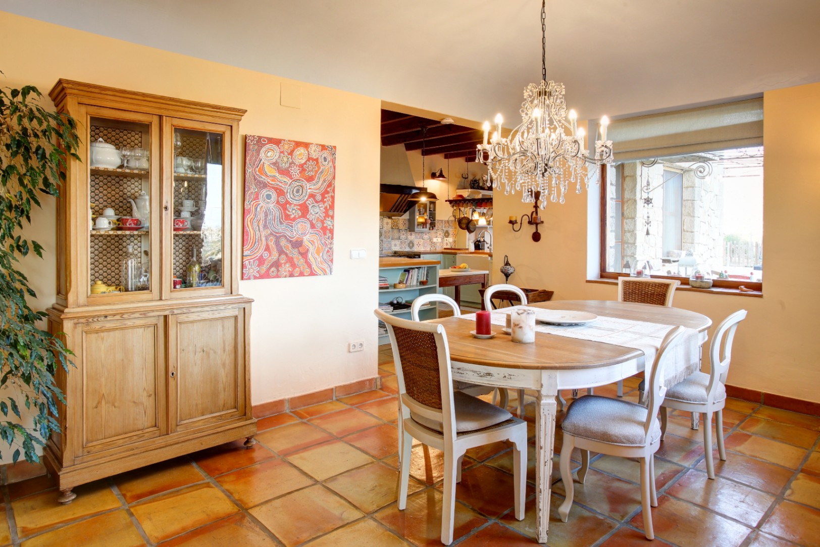 Eetkamer met houten tafel, witgekwaste stoelen en een kristallen kroonluchter, voorzien van een authentieke terracotta vloer en houten servieskast.