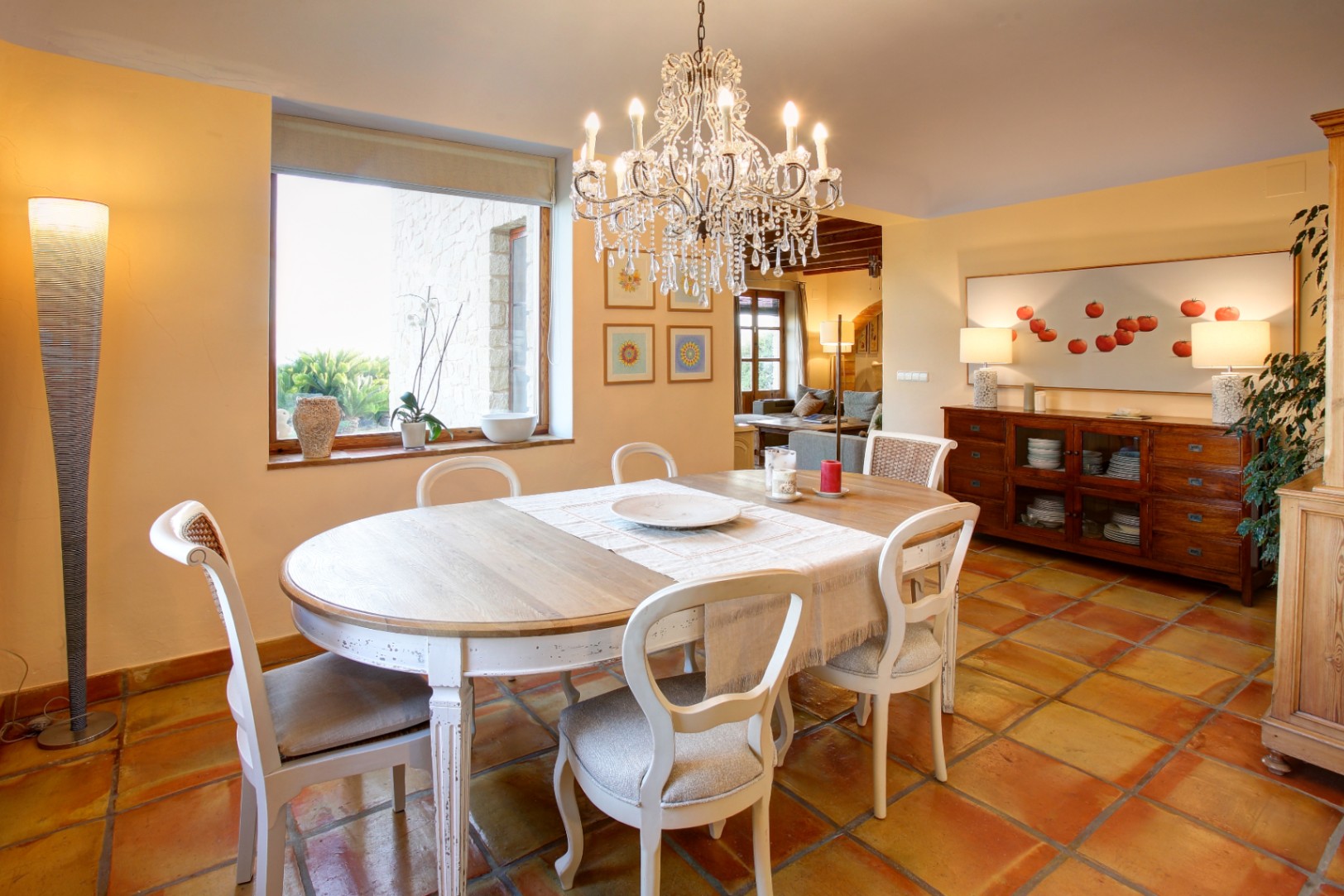 Eetkamer met terracotta vloer, een wit houten tafel en kristallen kroonluchter. Het grote raam zorgt voor natuurlijk licht en biedt uitzicht op de stenen buitenmuren.