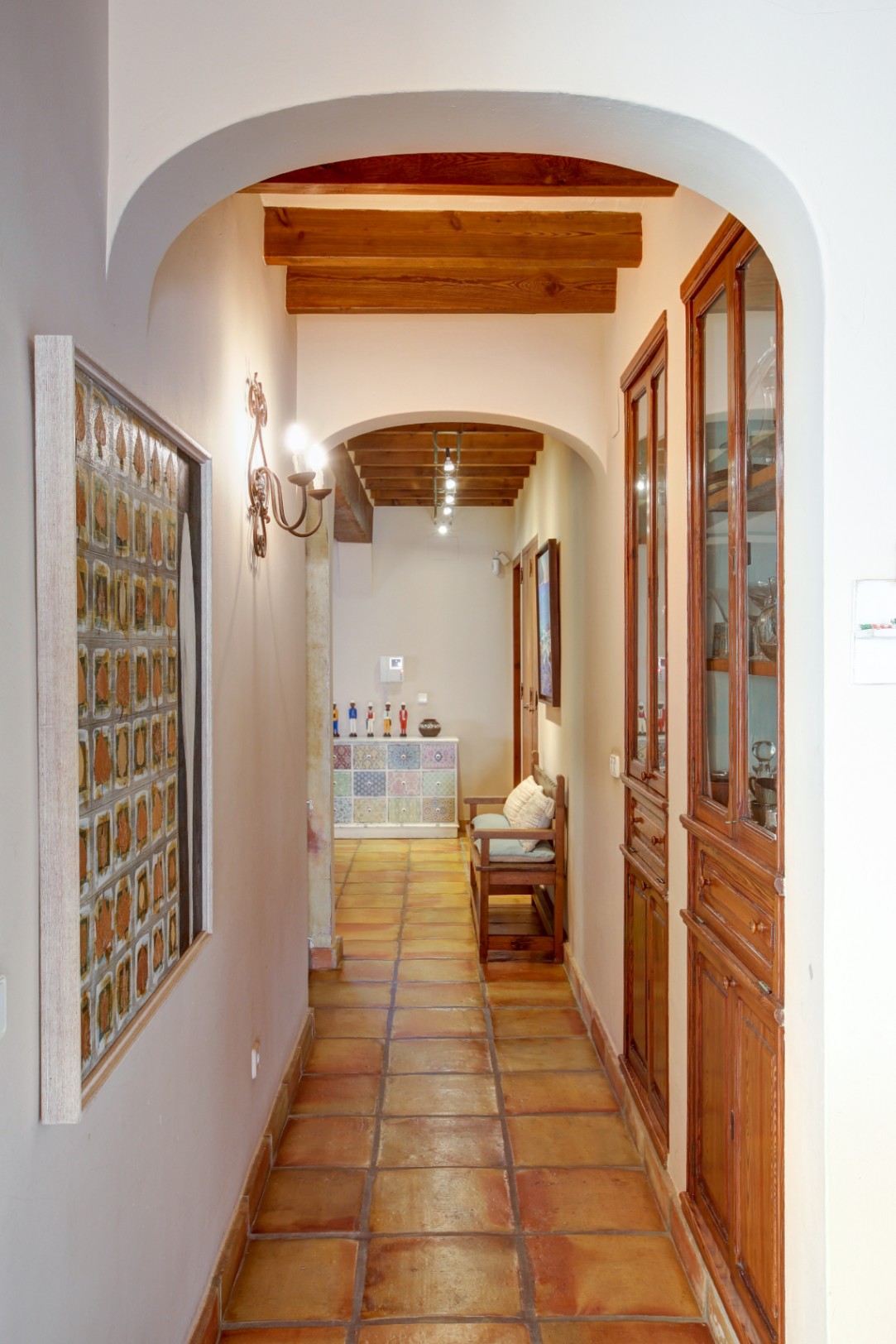 Gang met ronde bogen, zichtbare houten plafondbalken en een authentieke terracotta vloer. Voorzien van ingebouwde houten vitrinekasten en klassieke wandverlichting.