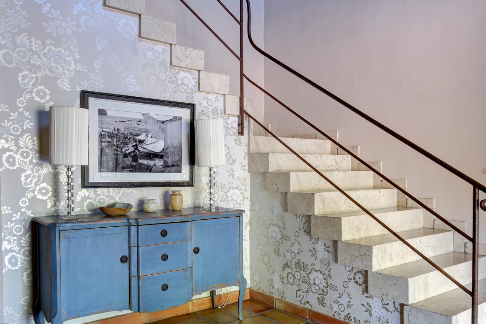 Massief marmeren trap met metalen leuning, naast een blauw gedecapeerd dressoir op een authentieke terracotta vloer met bloemenbehang.