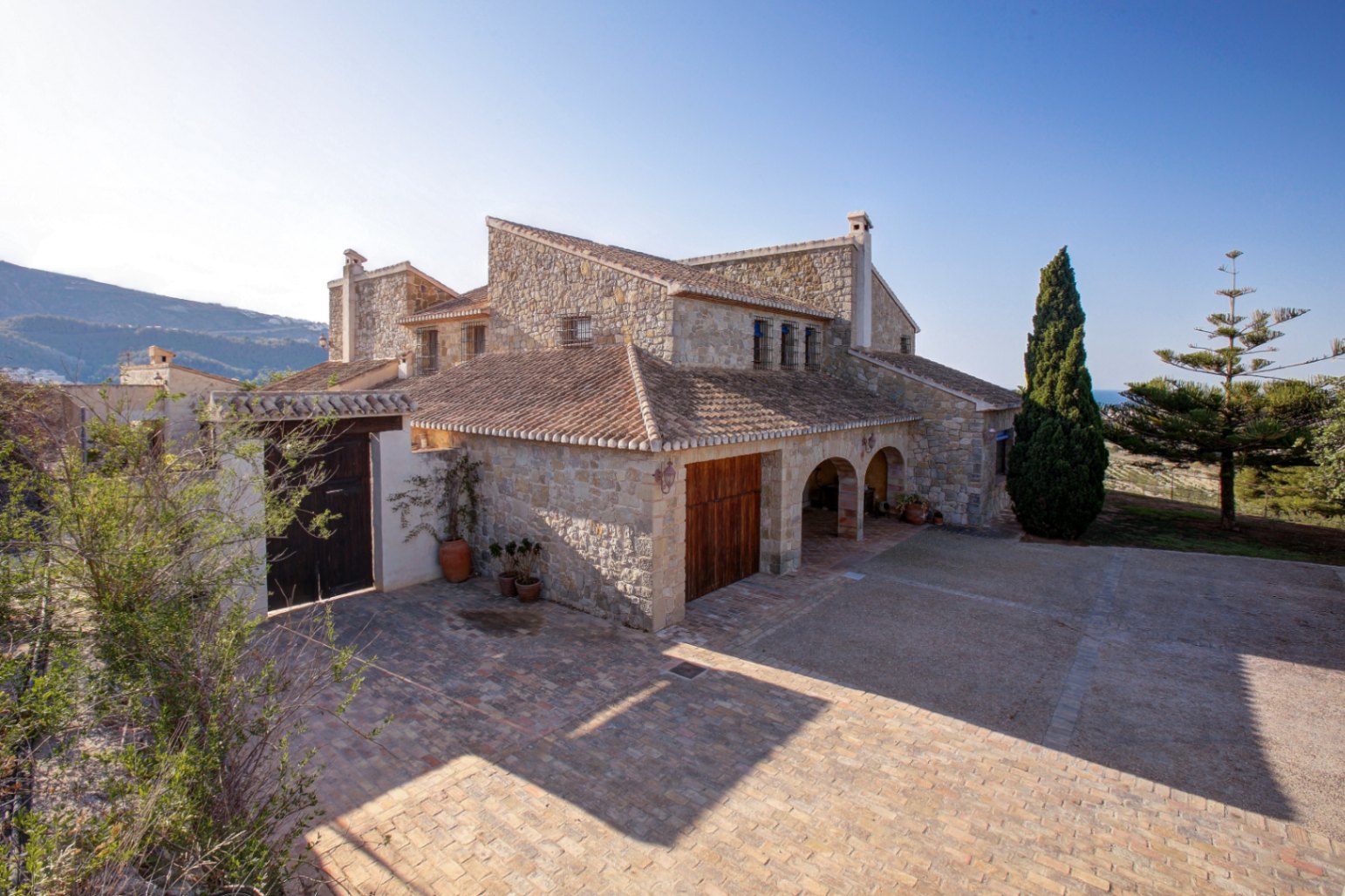 Natuurstenen gevel met traditionele bogen, keramische dakpannen en een ruim terras van gebakken klei onder de mediterrane zon.
