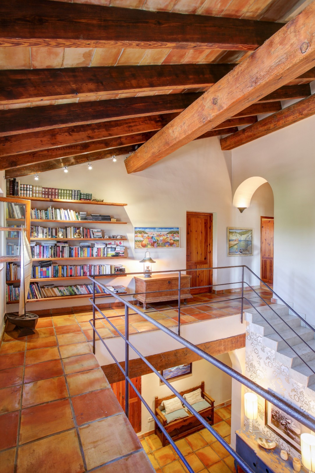 Overloop op de bovenverdieping met houten balkenplafond, terracotta vloertegels, een ingebouwde bibliotheek en uitzicht op de vide met smeedijzeren balustrade.