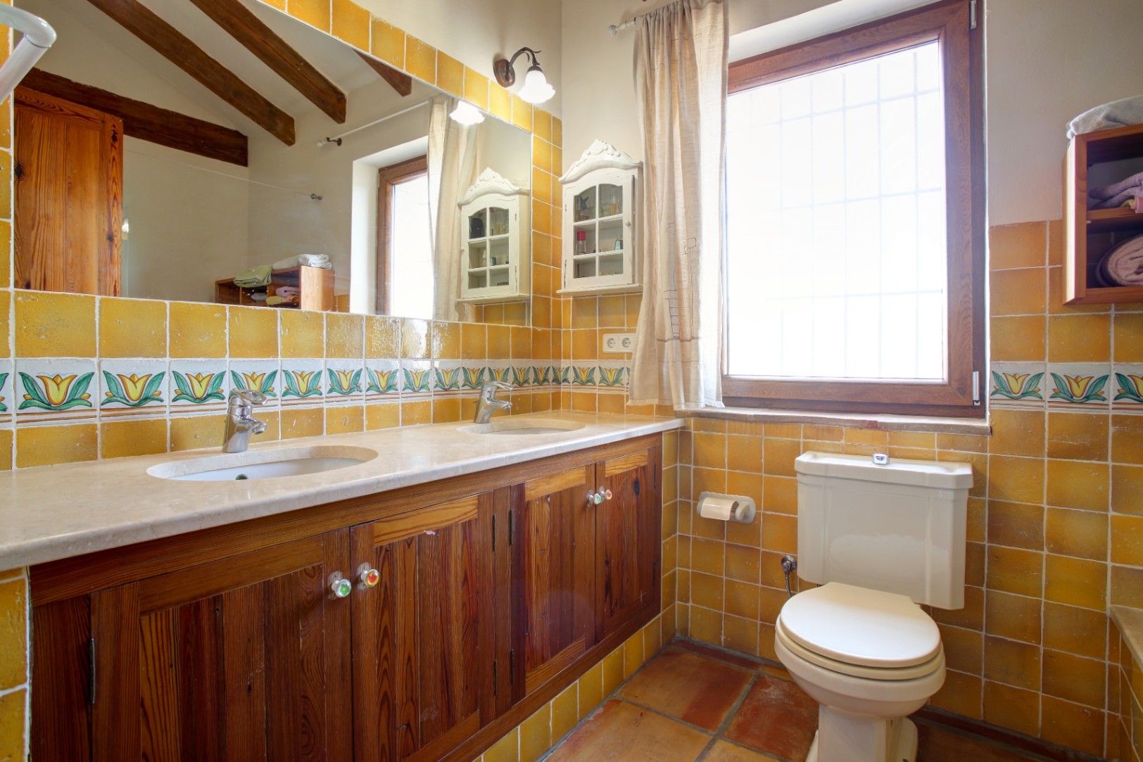 Rustieke badkamer met dubbele marmeren wastafel, houten kasten en mosterdgele tegels. Voorzien van zichtbare houten balken en een traditionele terracotta vloer.