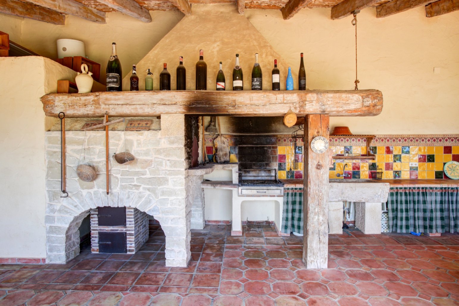 Rustieke buitenkeuken met ingebouwde barbecue, massieve houten balk, natuurstenen gootsteen en kleurrijk tegelwerk op een authentieke terracotta vloer.