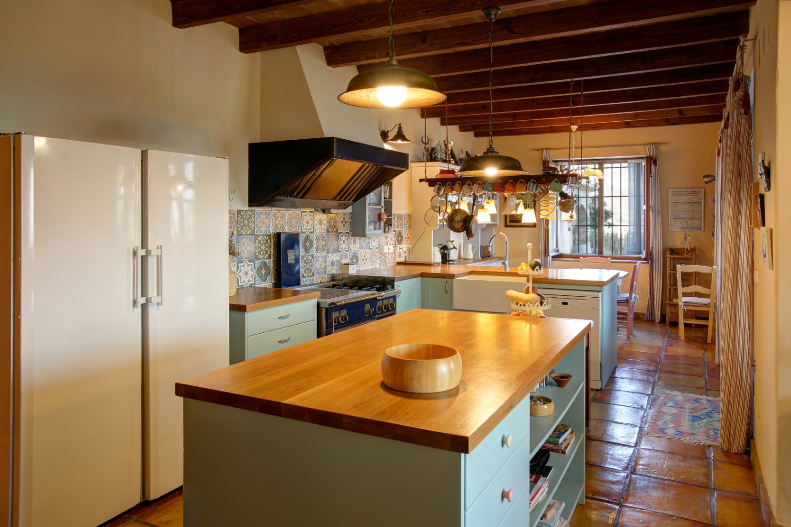 Rustieke keuken met houten kookeiland, zichtbare balken en terracotta vloer. Voorzien van een blauw professioneel fornuis, decoratieve tegelwand en natuurlijke lichtinval.