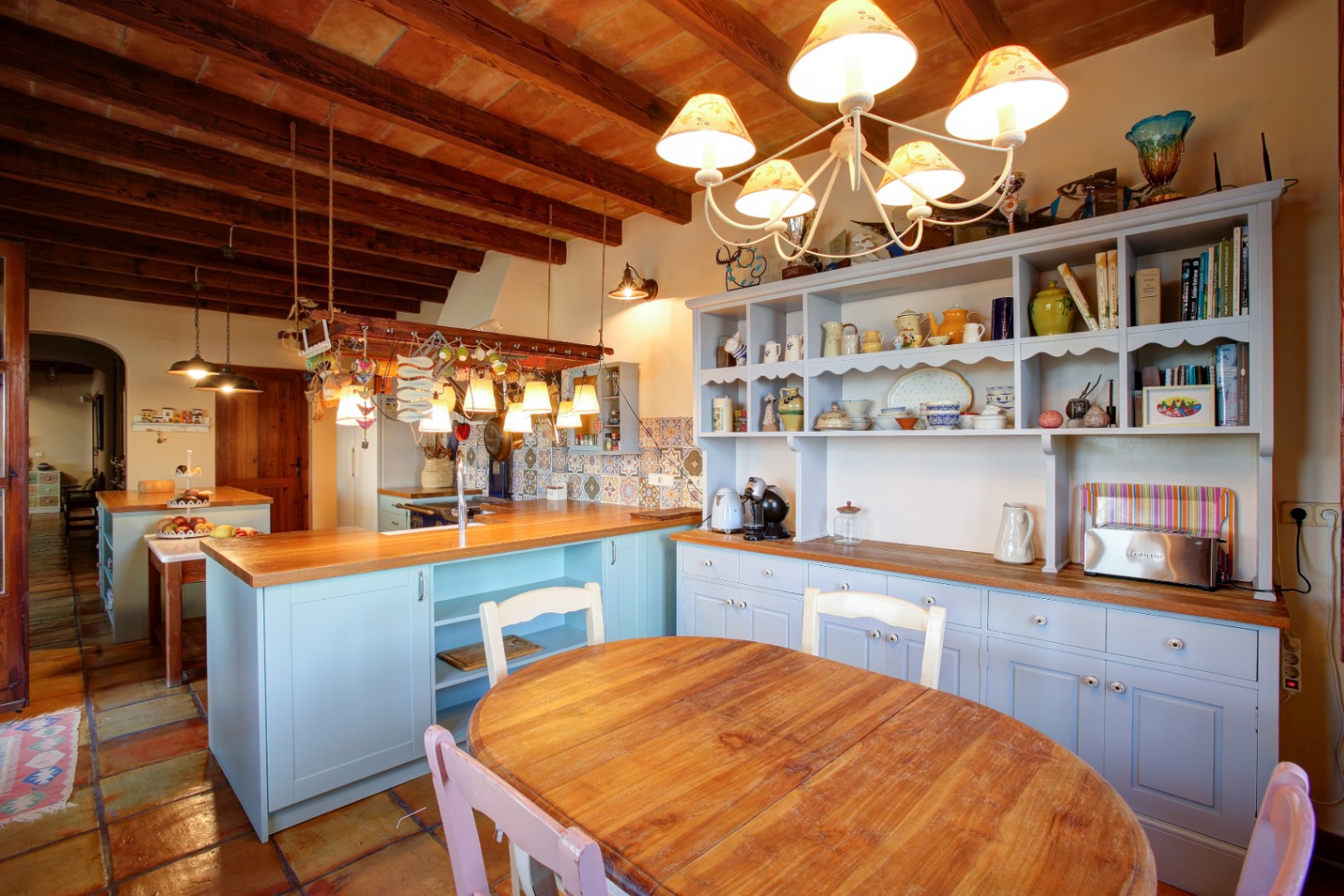 Rustieke keuken met lichtblauwe kasten, houten werkbladen en een balkenplafond. De ruimte is voorzien van een eethoek met authentieke terracotta vloertegels.