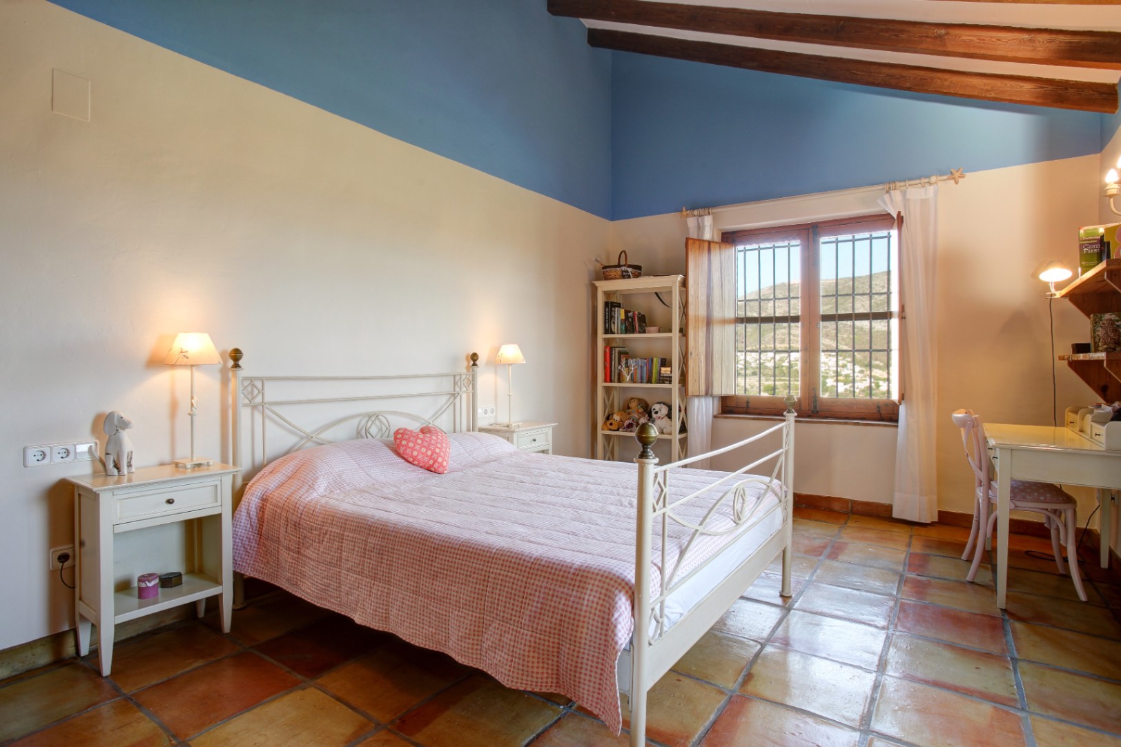 Rustieke slaapkamer met houten balkenplafond, terracotta vloertegels en een wit smeedijzeren bed. De kamer heeft een bureau en uitzicht op de omliggende heuvels.