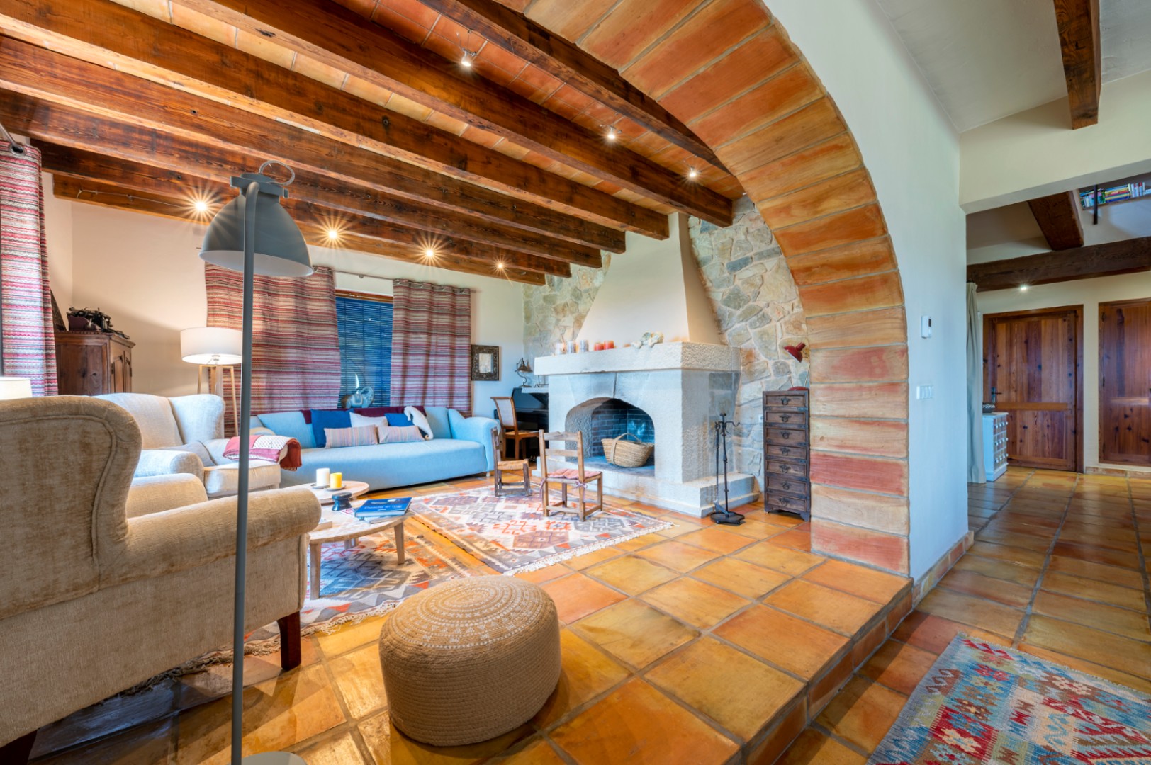 Rustieke woonkamer met stenen open haard, plafond met houten balken en terracotta tegels, gebakken kleivloeren en een karakteristieke bakstenen boog.