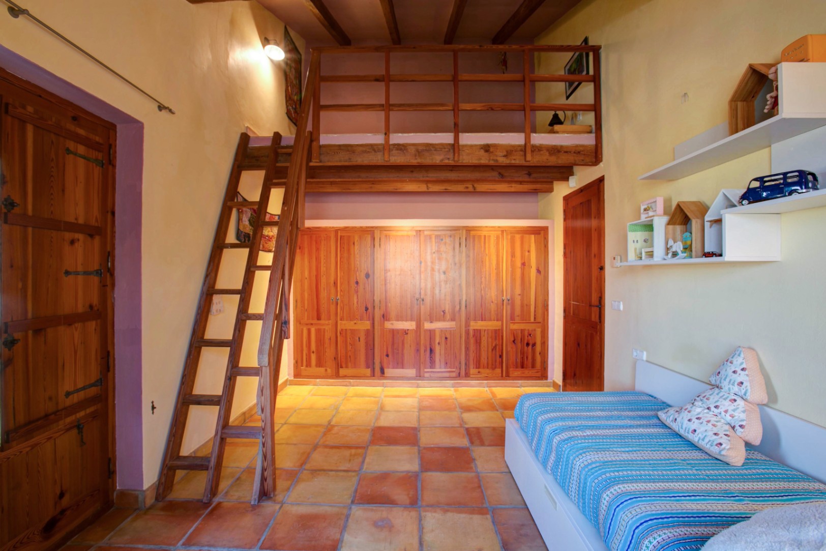 Slaapkamer met houten entresol en ladder, ingebouwde kasten van mobila-hout en traditionele terracotta tegelvloer onder zichtbare balken.