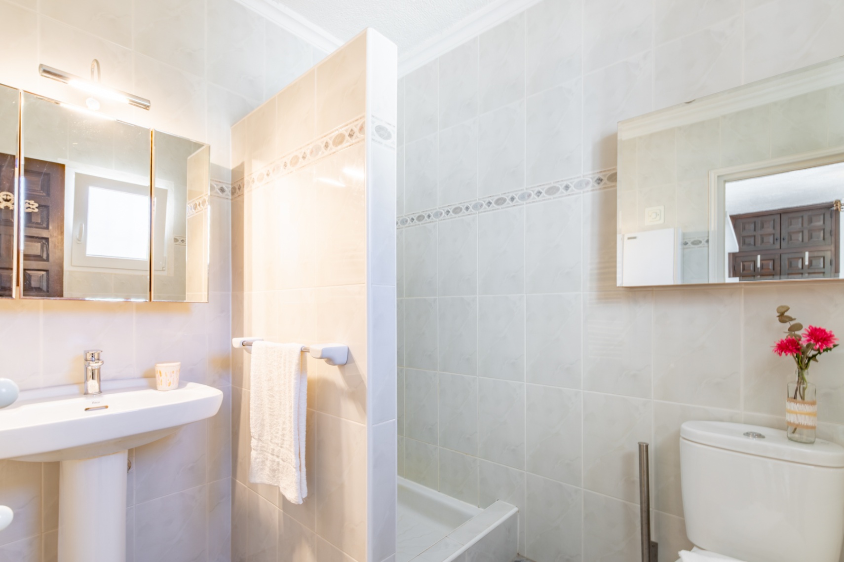 Badkamer met crème tegels, een wastafel op zuil, spiegelkast met verlichting en een doucheruimte gescheiden door een betegelde wand.