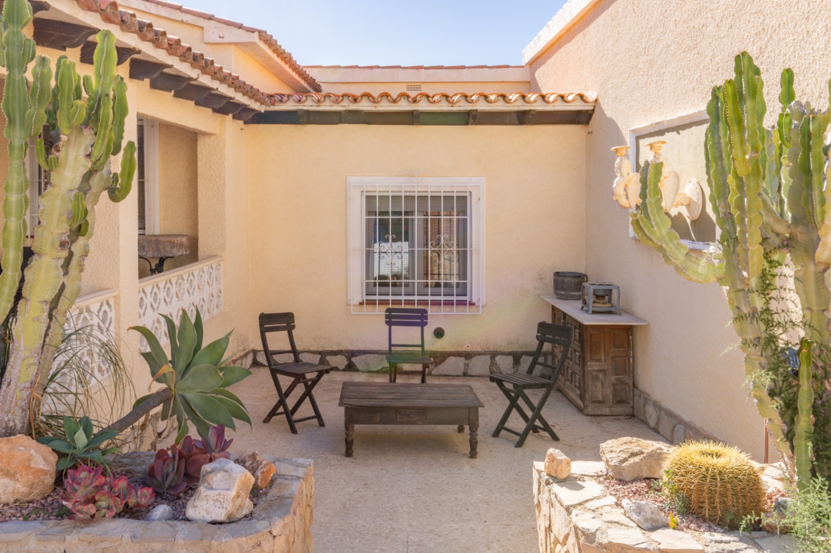 Buitenpatio in mediterrane stijl met stenen vloer, rustiek houten meubilair en stenen plantenbakken met cactussen en vetplanten in de middagzon.