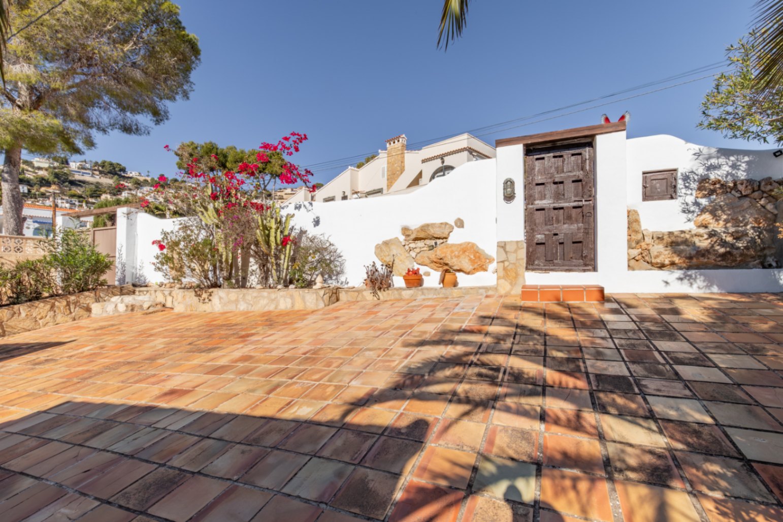 Buitenterras met terracotta tegels, witte muren met natuurstenen accenten en een rustieke houten poort omgeven door mediterrane bougainville en cactussen.