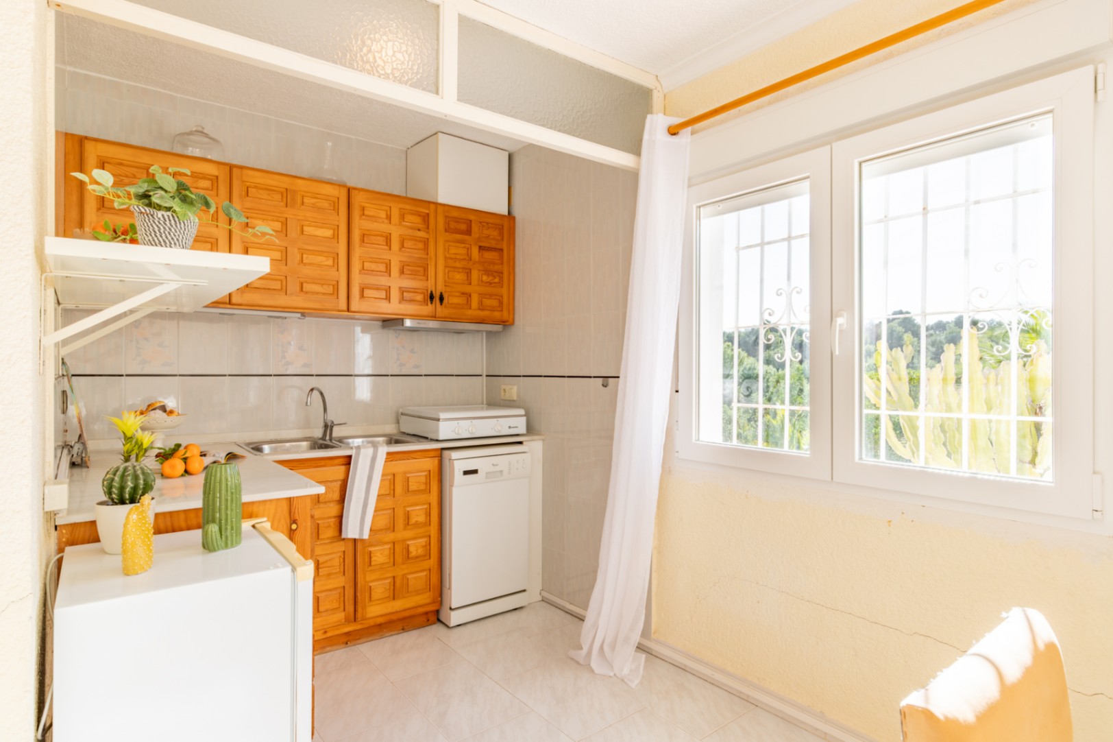 Functionele kitchenette van het onafhankelijke appartement met houten kasten, wit werkblad en een groot raam met overvloedig natuurlijk licht.