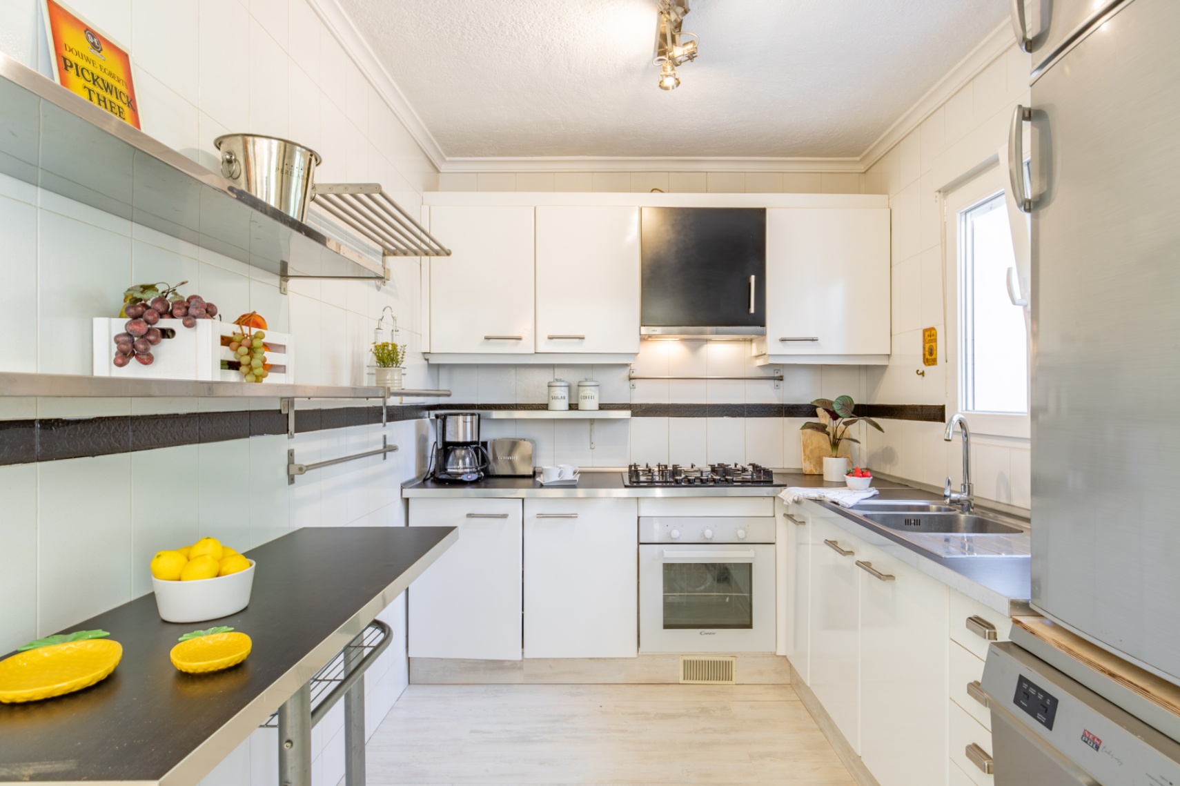 Ingerichte keuken met witte kasten, donkere werkbladen, roestvrijstalen rekken en natuurlijk licht. Voorzien van gaskookplaat, oven en dubbele spoelbak.