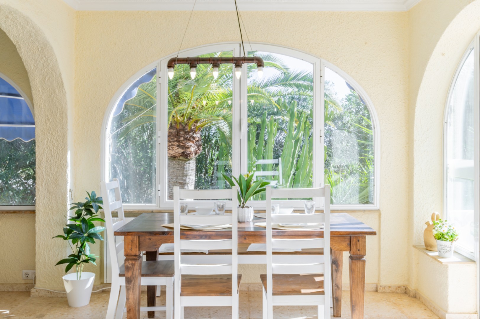 Lichte eethoek in een glazen naya met houten tafel, witte stoelen en industriële verlichting, uitkijkend op de tropische tuin met palmen.