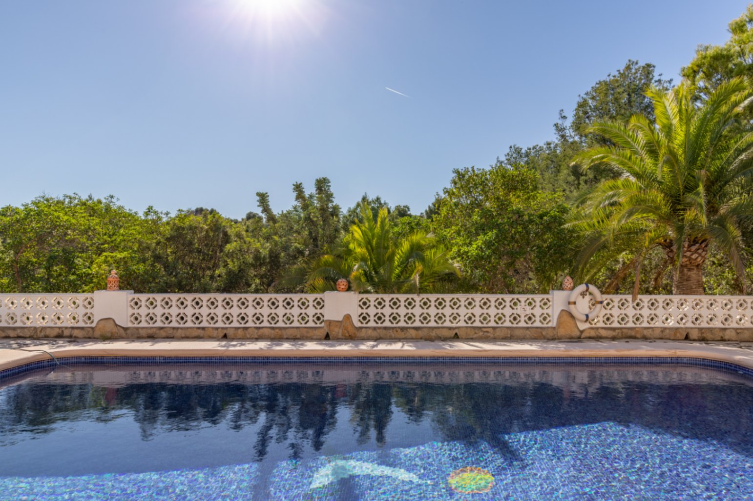 Op het zuiden gelegen zwembad met blauwe mozaïektegels, afgezet met een witte decoratieve balustrade en omringd door volwassen mediterrane beplanting.