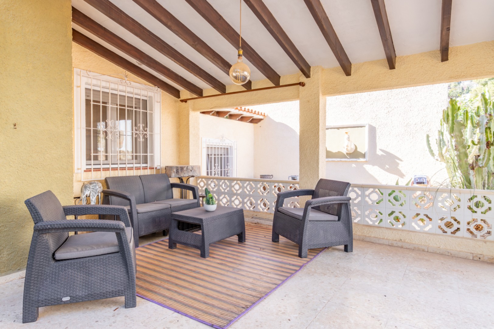 Overdekt terras met houten balkenplafond, grijze wicker tuinmeubelen en een decoratieve balustrade met uitzicht op de mediterrane tuin.