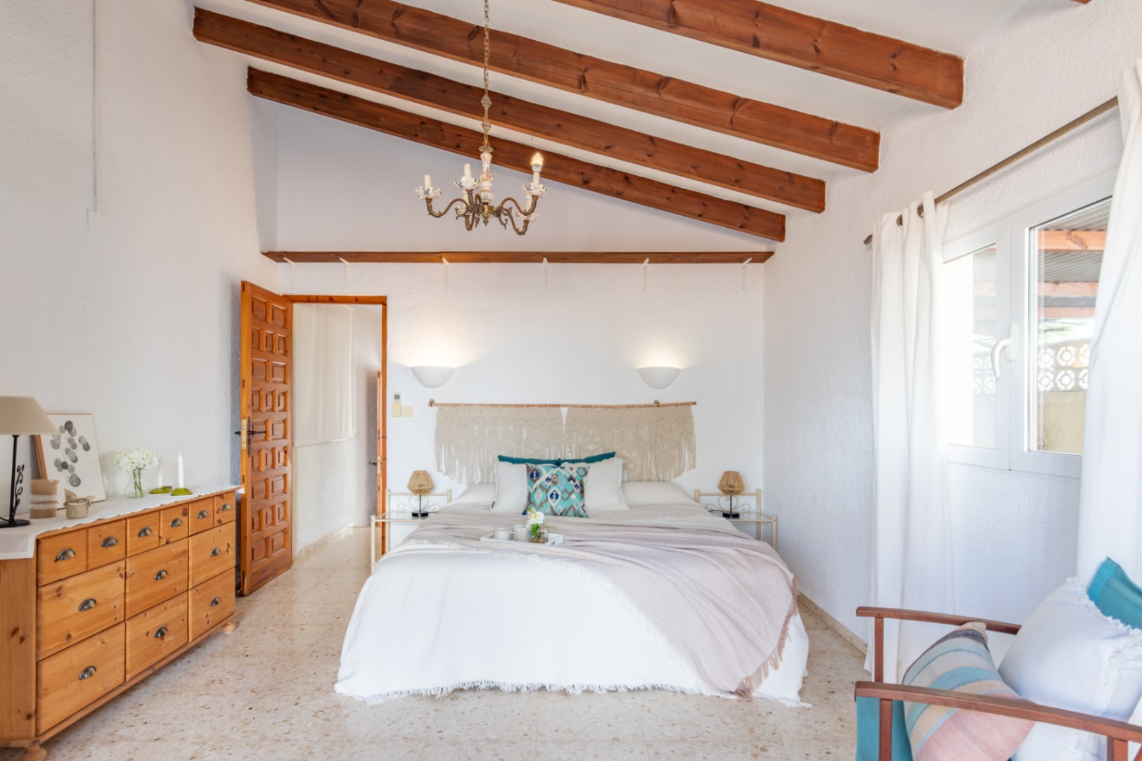 Slaapkamer met hoge plafonds met houten balken, een terrazzovloer en mediterrane decoratie met een macramé hoofdbord en houten meubilair.