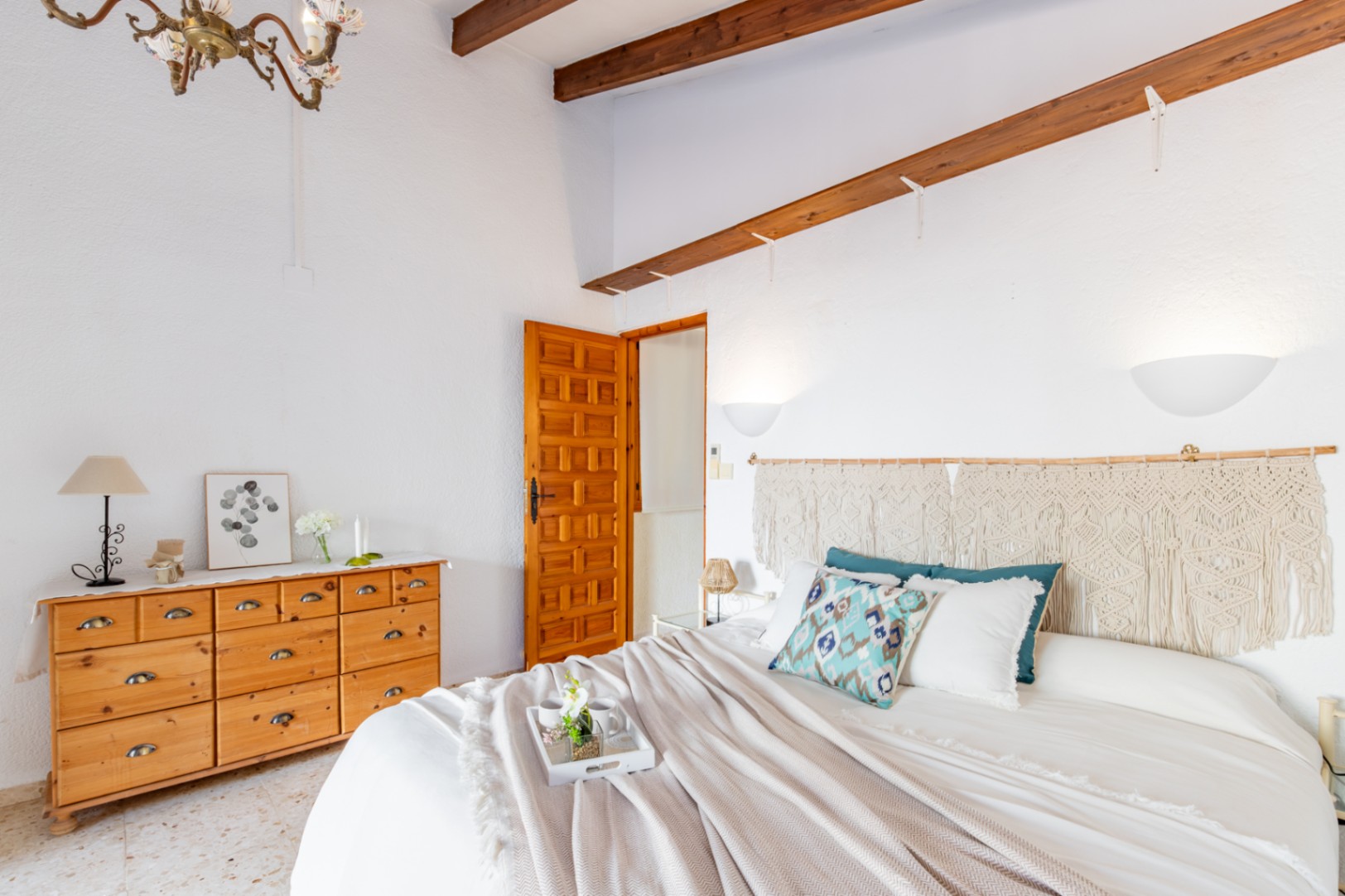Slaapkamer met houten plafondbalken, een grenen ladenkast en een macramé hoofdbord. Voorzien van een originele terrazzovloer en een traditionele bewerkte houten deur.