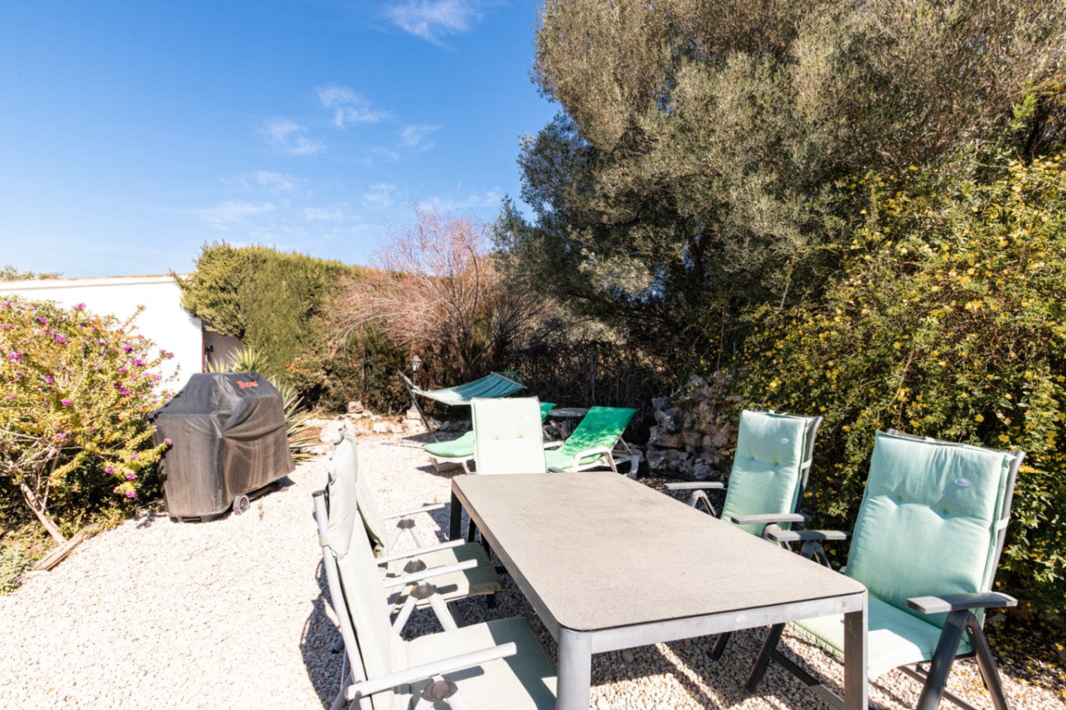 Buiteneethoek op grind met een rechthoekige tafel, gevoerde stoelen, een afgedekte barbecue en een hangmat tussen de volgroeide mediterrane beplanting.