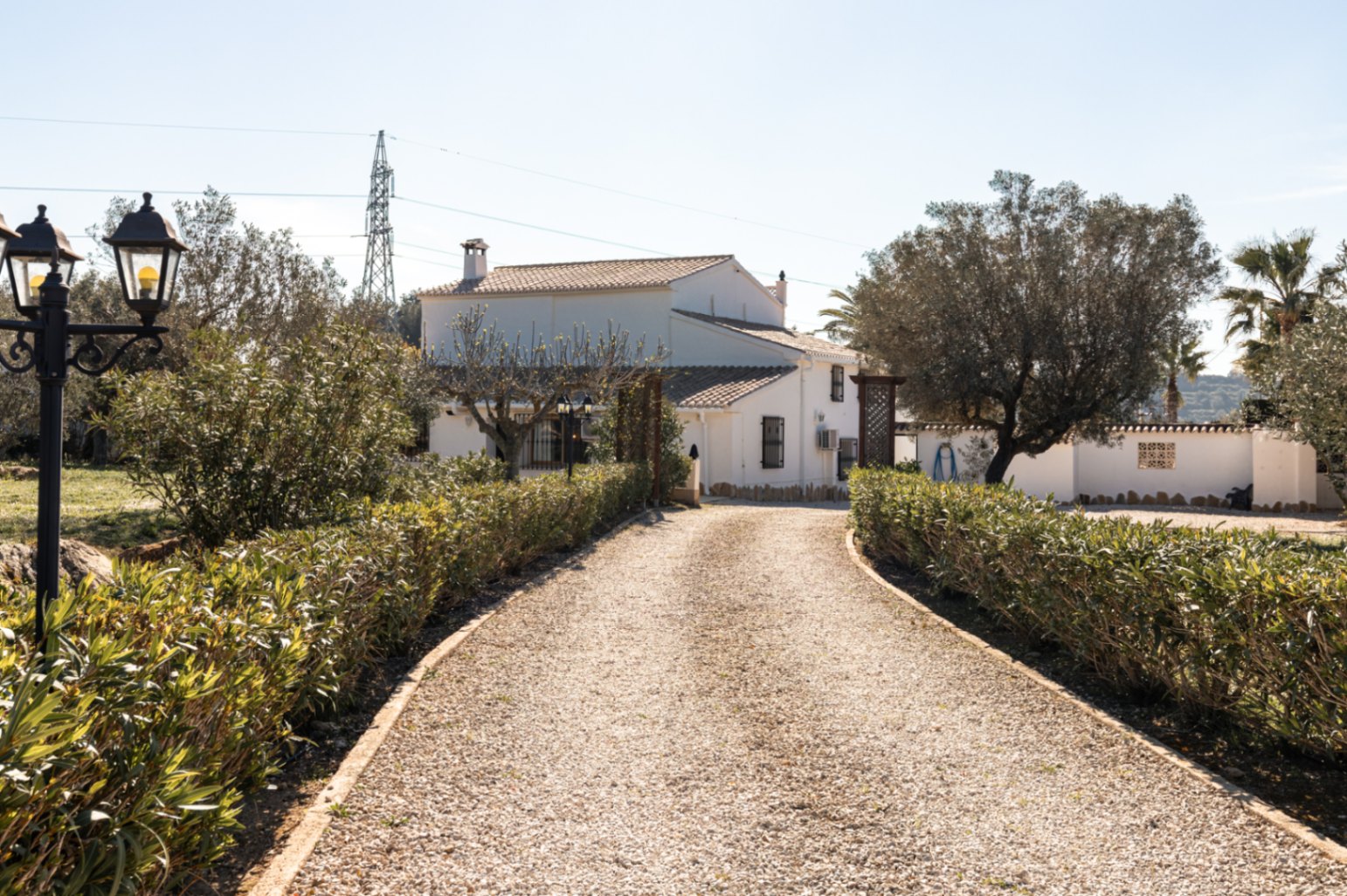 Een grindpad geflankeerd door lage hagen leidt naar de witgekalkte gevel van de finca, met traditionele mediterrane architectuur en volgroeide bomen.