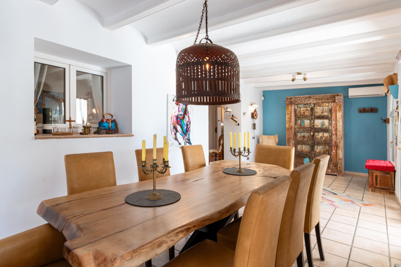 Eethoek met een massief houten tafel met natuurlijke rand, gestoffeerde stoelen en wit balkenplafond, geaccentueerd door een blauwe muur en rustieke elementen.