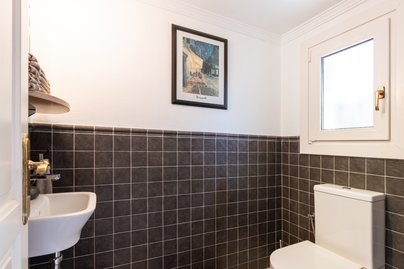 Functioneel gastentoilet met donkere getextureerde wandtegels, een compacte wastafel en een draai-kiepraam voor natuurlijke ventilatie.