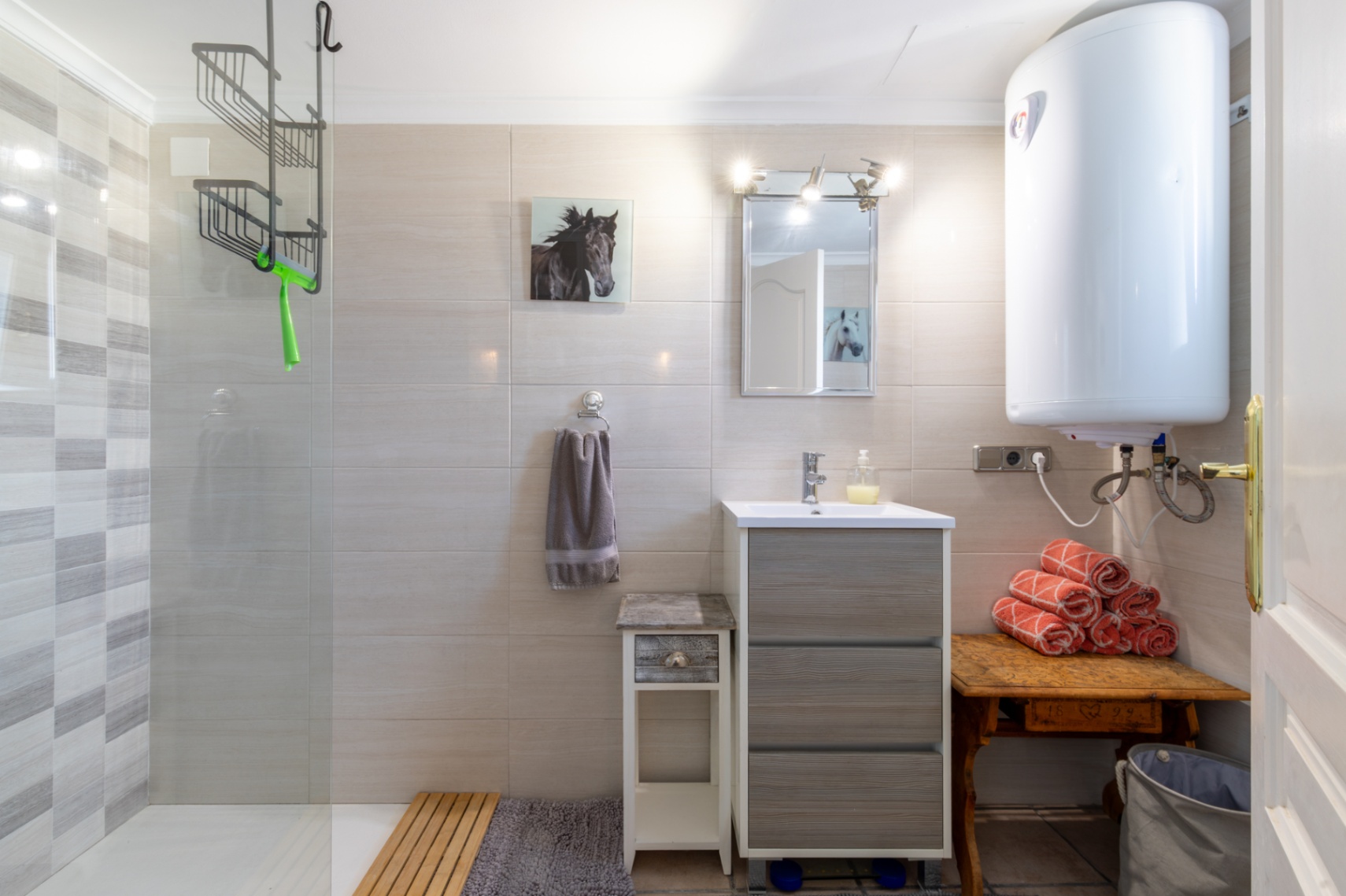 Functionele badkamer met inloopdouche, glazen wand en getextureerde tegels. Voorzien van een grijs wastafelmeubel, elektrische boiler en rustieke houten accenten.