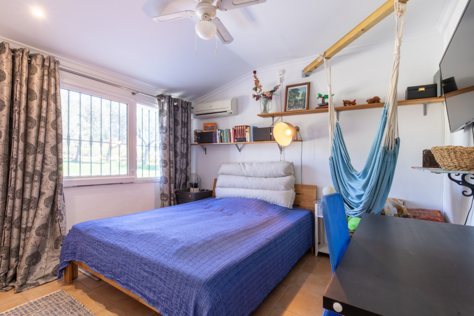 Functionele slaapkamer met tweepersoonsbed, bureau, houten planken en een hangmat. Voorzien van airconditioning, plafondventilator en veel natuurlijk lichtinval.