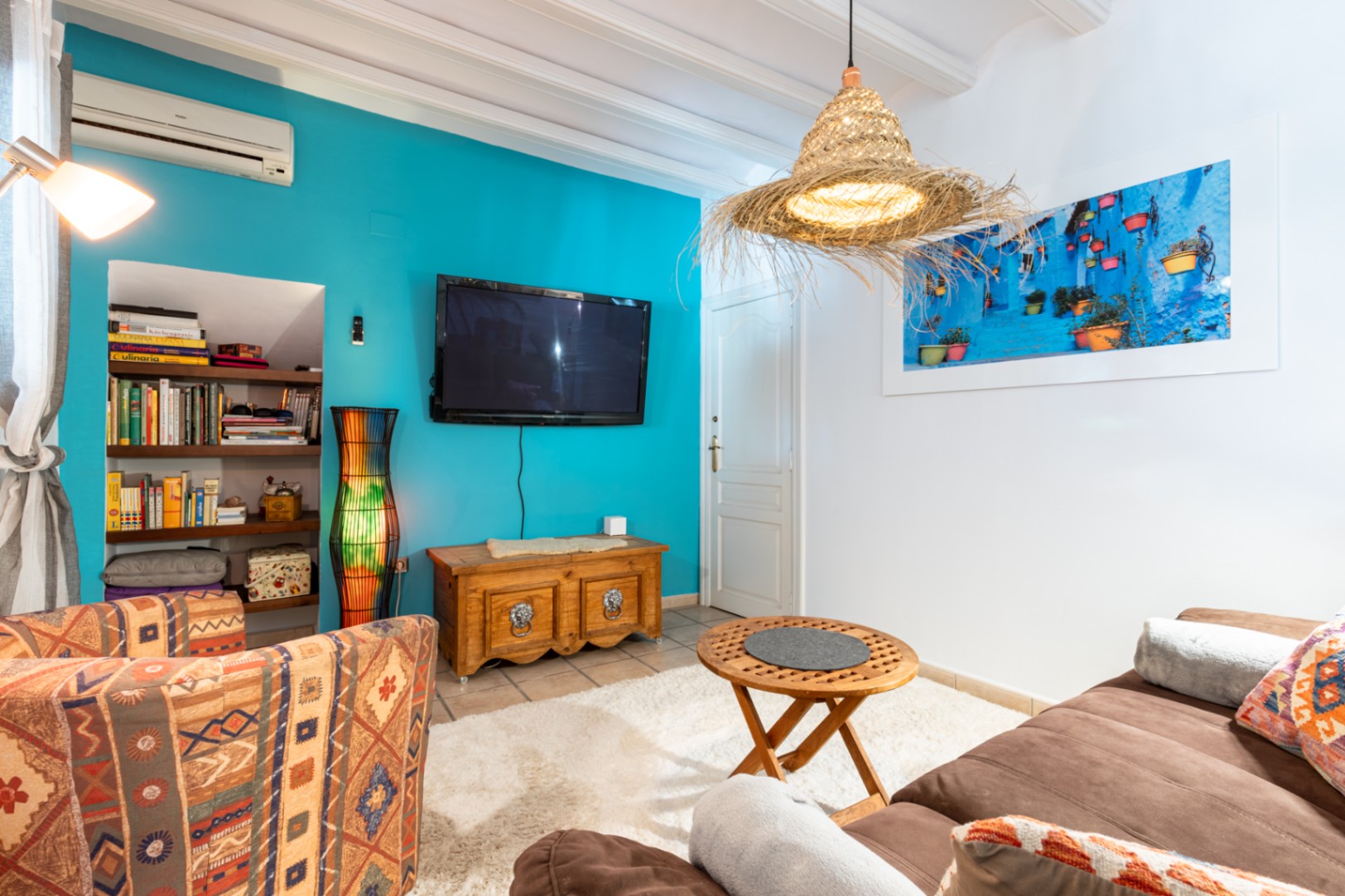 Gezellige woonkamer met een turquoise accentmuur, wit balkenplafond en ingebouwde boekenkast. Voorzien van airconditioning, rustieke houten meubels en een natuurlijke hanglamp.