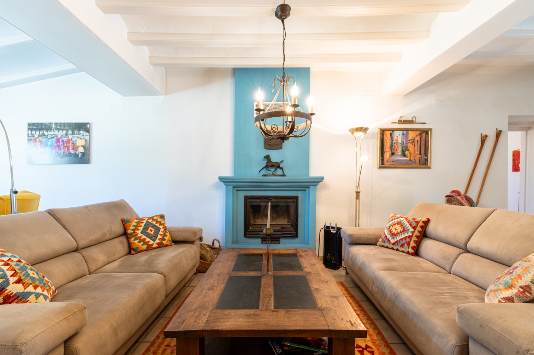 Lichte woonkamer met wit balkenplafond, een opvallende blauwe haard en comfortabele banken rond een robuuste salontafel van hout en leisteen.