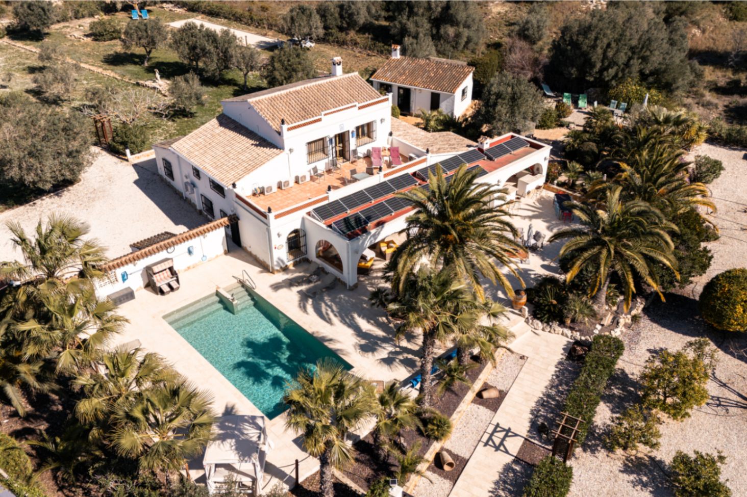 Luchtfoto van de finca met het rechthoekige zwembad, stenen terrassen en zonnepanelen op de naya, omgeven door een volgroeide mediterrane tuin.