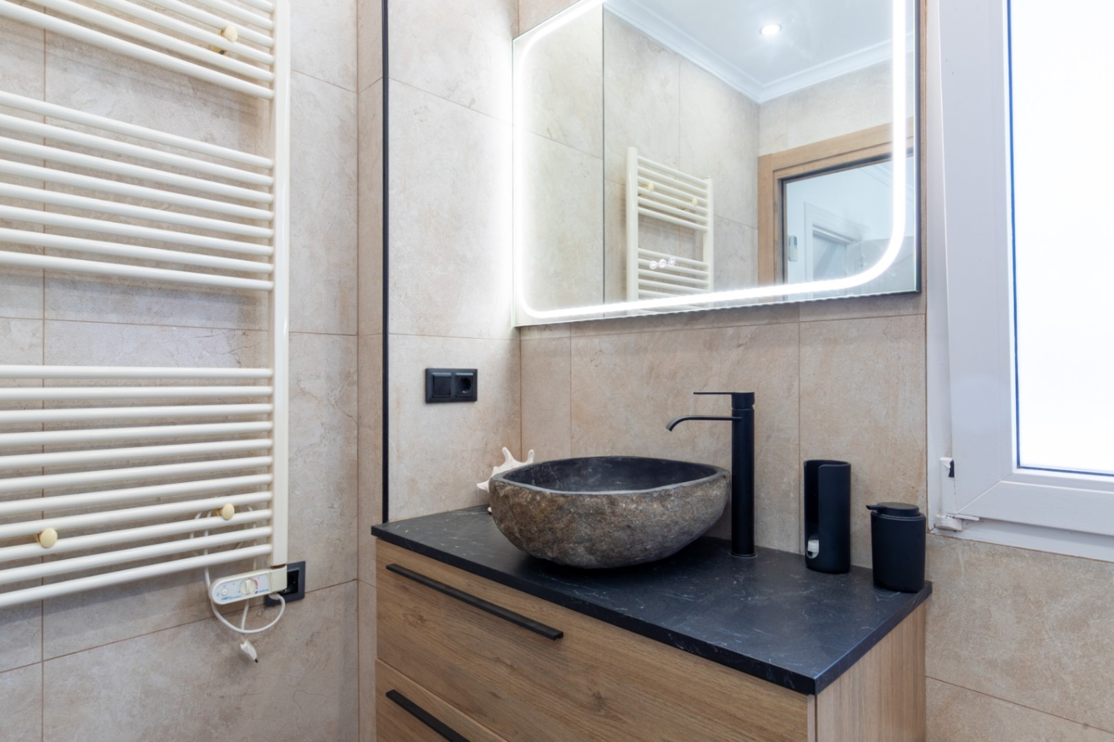 Moderne badkamer met een wastafel van natuursteen op een zwart werkblad en houten meubel. Voorzien van een LED-verlichte spiegel en een witte handdoekradiator.