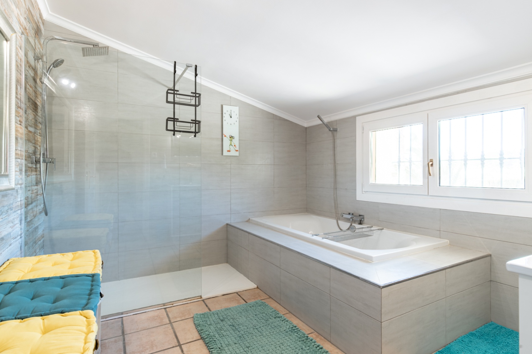 Moderne badkamer met inloopdouche, glazen wand en ingebouwd ligbad. Voorzien van grijze wandtegels, chromen kranen en natuurlijke lichtinval via het raam.