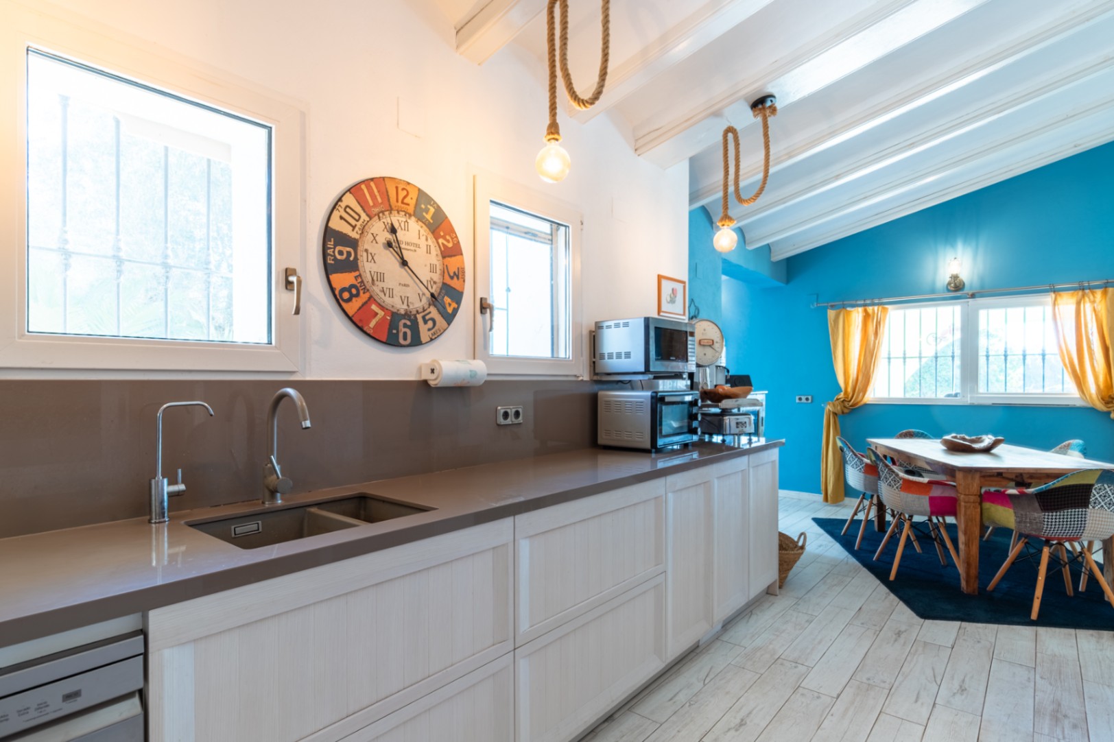 Moderne keuken met kwarts werkblad, open naar een lichte eethoek met blauwe accentmuur, witte balkenplafonds en rustieke touwverlichting.