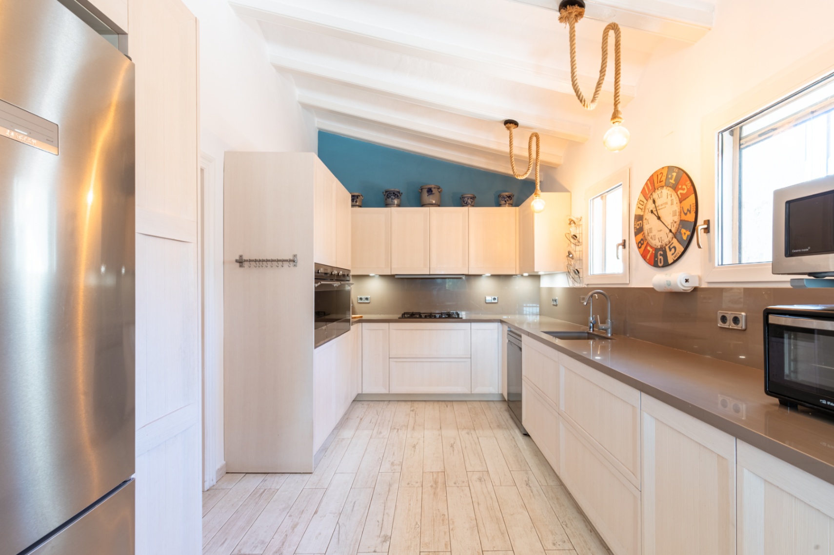 Moderne keuken met stenen werkbladen, rvs-apparatuur en een wit balkenplafond. Voorzien van decoratieve touwverlichting en een blauwe accentmuur boven de kasten.