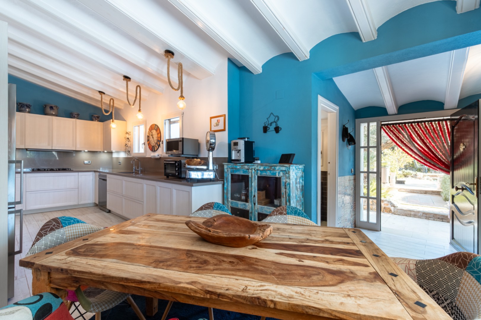 Moderne keuken met witte balkenplafonds, blauwe muren, een rustieke houten tafel en touwverlichting. Voorzien van hoogwaardige apparatuur en directe toegang naar buiten.