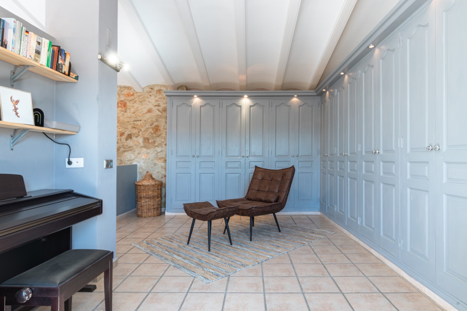 Overloop met uitgebreide ingebouwde kasten in zachtblauw, natuurstenen muur, wit balkenplafond en een zithoek met leren fauteuil en piano.