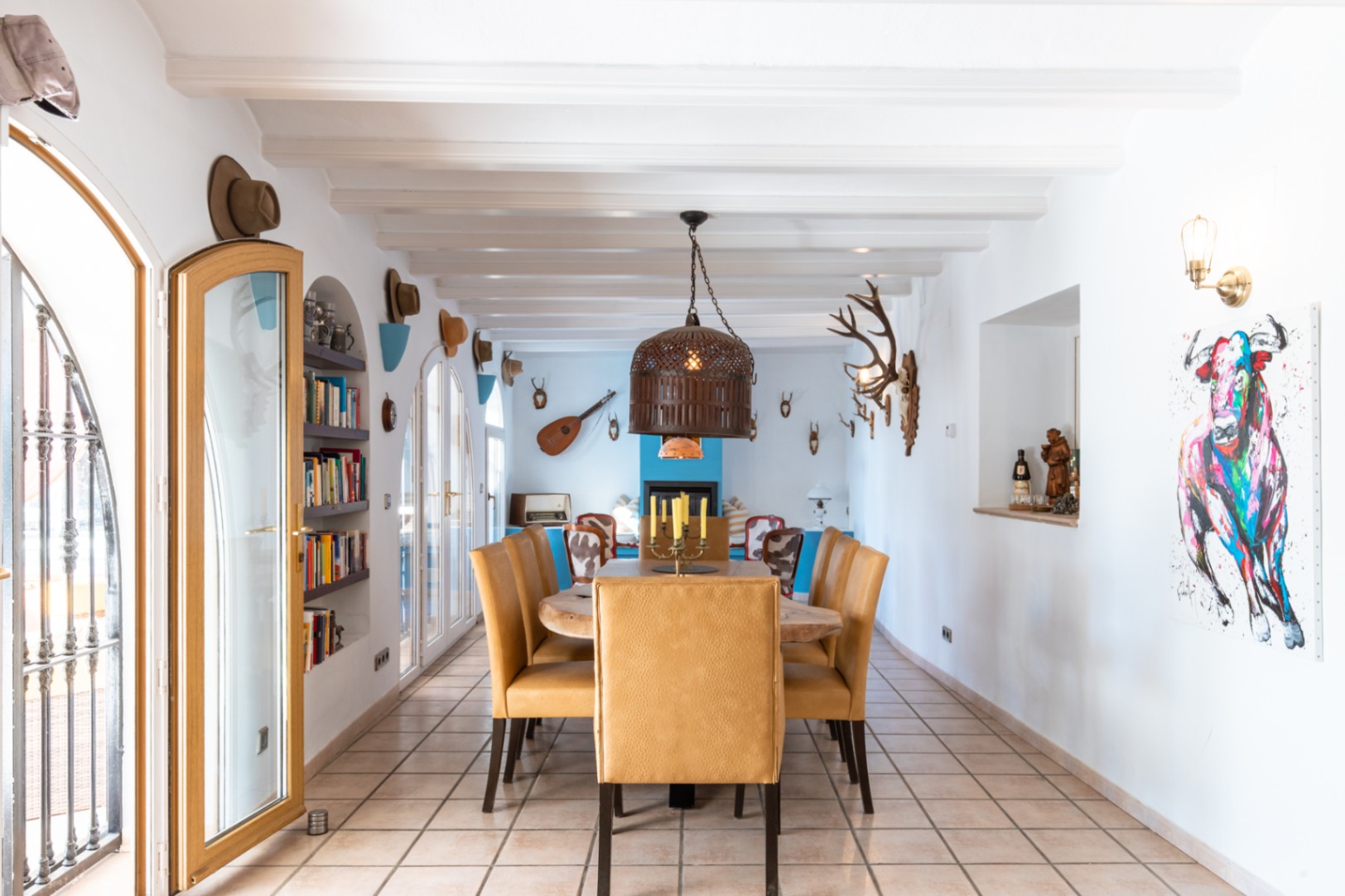 Ruime eetkamer met witte balkenplafonds, een massief houten tafel en mosterdgele stoelen. De ruimte heeft karakteristieke boogdeuren, ingebouwde boekenkasten en een rustieke afwerking.