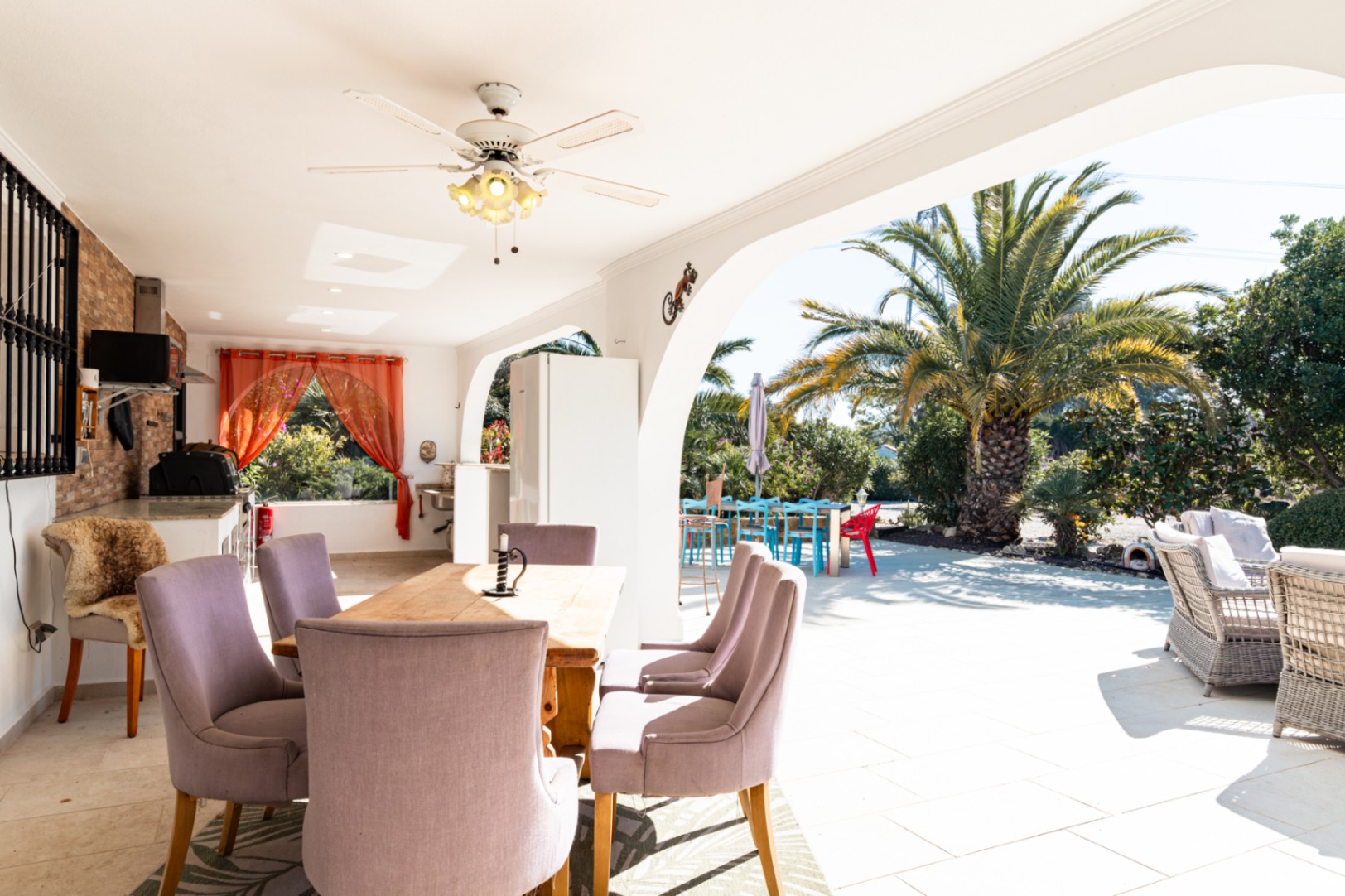 Ruime overdekte naya met eethoek, plafondventilator en een buitenkeuken. Het terras kijkt uit op een zonnige tuin met palmbomen en comfortabel buitenmeubilair.