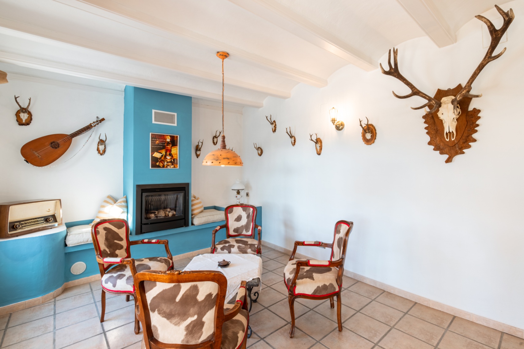 Rustieke zithoek met een gashaard in een blauwe wand, fauteuils bekleed met koeienhuid en een wit balkenplafond gedecoreerd met jachttrofeeën.