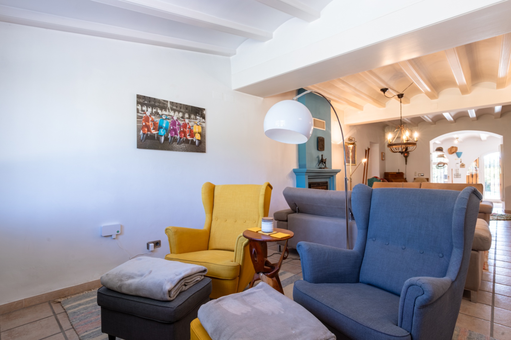 Sfeervolle woonkamer met wit balkenplafond, blauwe schouw en kleurrijke oorfauteuils onder een moderne booglamp.