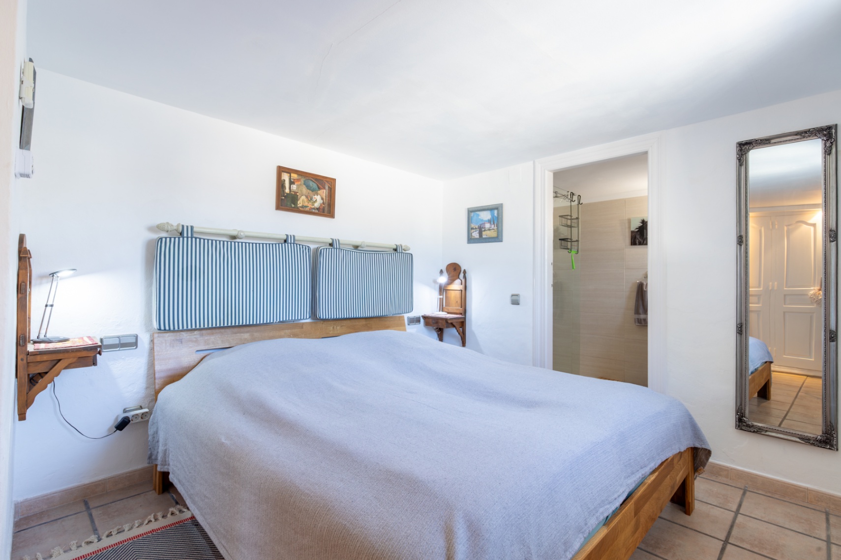 Slaapkamer met houten bedframe, gestreepte hoofdbordkussens en wandgemonteerde nachtkastjes. Inclusief directe toegang tot de en-suite badkamer en een grote sierlijke wandspiegel.