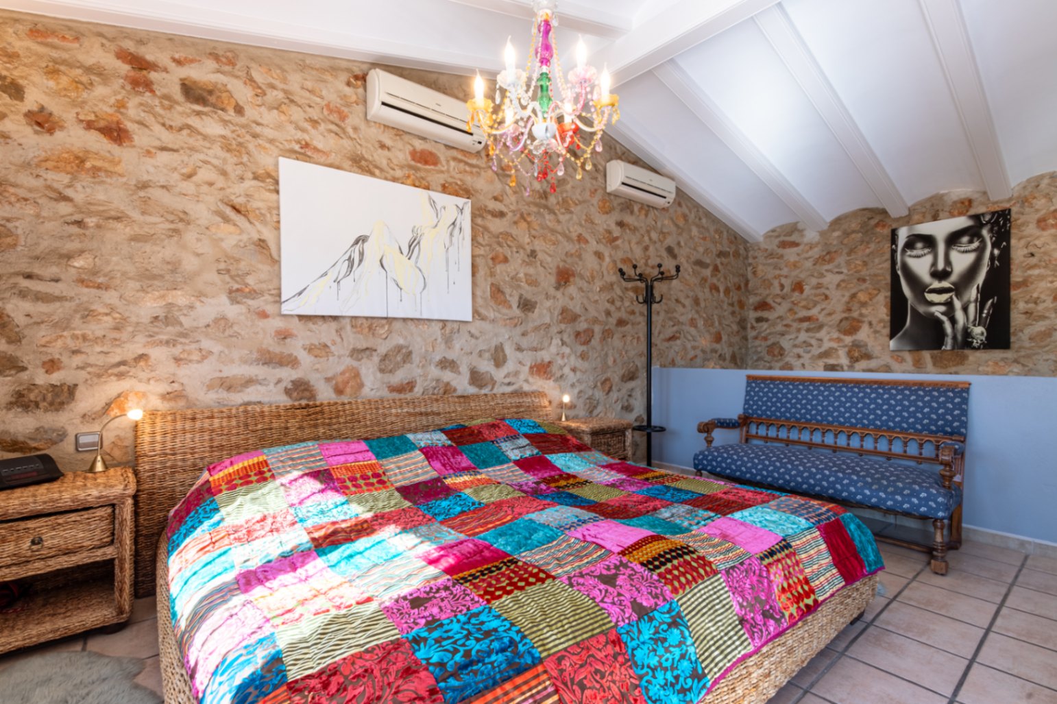 Slaapkamer met natuurstenen muren, een wit balkenplafond en een rieten bed met een kleurrijke patchwork sprei onder een kristallen kroonluchter.